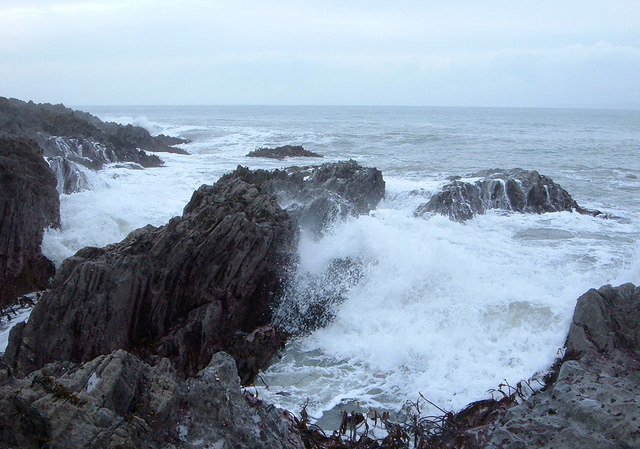 Rocky coastline with crashing waves under an overcast sky. The turbulent sea is white with foam, conveying a sense of raw power and rugged beauty.