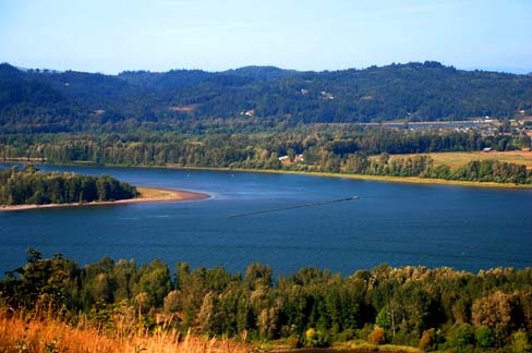 Wide river bends through lush forest and fields under a clear blue sky. Hills in the distance create a serene, peaceful landscape.