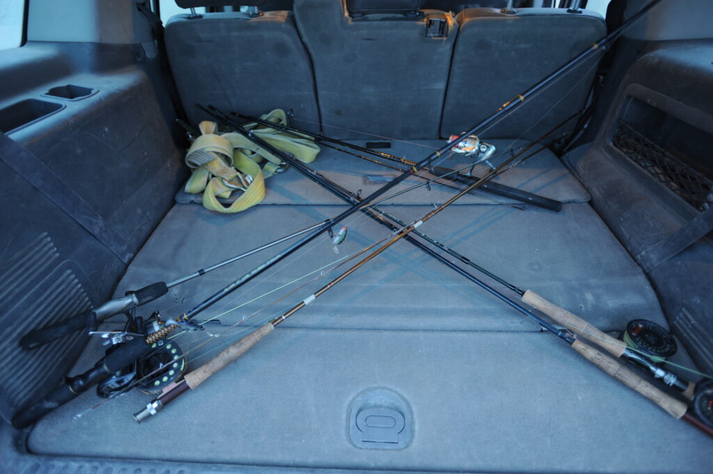 A collection of fishing rods with reels lies crossed in the back of a car, alongside coiled yellow straps. The setting suggests an upcoming fishing trip.