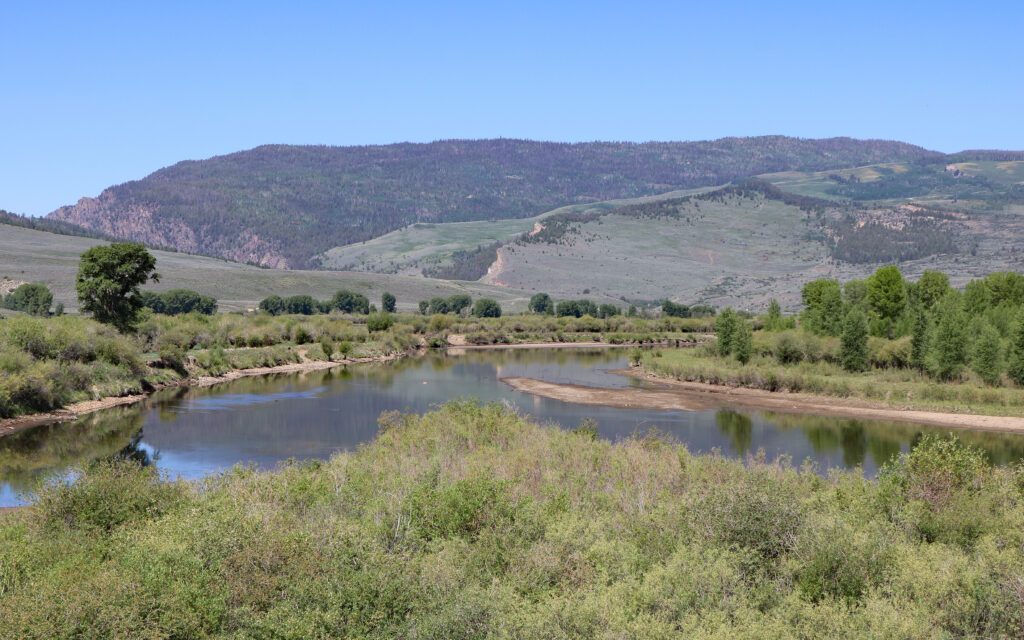 A serene landscape with a winding river reflecting the clear blue sky, surrounded by lush greenery and distant rolling mountains in the background.