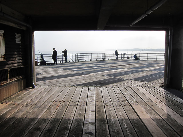 People fish on a wooden pier, silhouetted against a bright, hazy sky. The foreground shows wet planks, creating a serene and tranquil scene.
