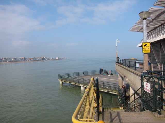 Coastal view from a pier showing calm sea and a distant shoreline under a clear sky. A CCTV sign is visible, conveying a sense of surveillance.