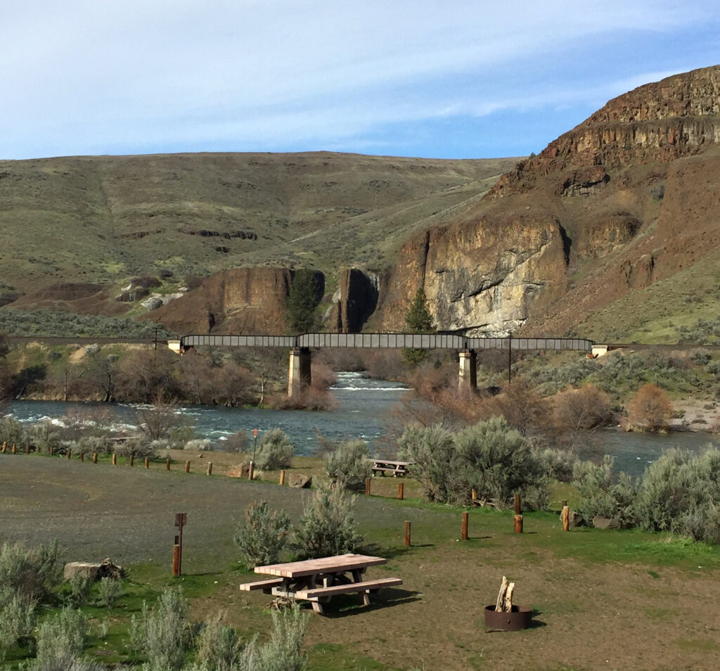 A scenic landscape with a tranquil river flowing under a bridge, surrounded by rocky hills and green shrubs. A picnic table and fire pit rest in the foreground, conveying a peaceful, inviting atmosphere.