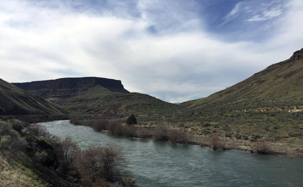 A serene river flows between green hills under a partly cloudy sky, with a flat-topped mountain in the background, evoking a peaceful, natural scene.
