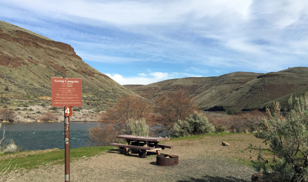 Campsite with picnic table and fire ring near a river, surrounded by desert hills and shrubs under a blue sky with wispy clouds. Sign reads "Group Campsite."