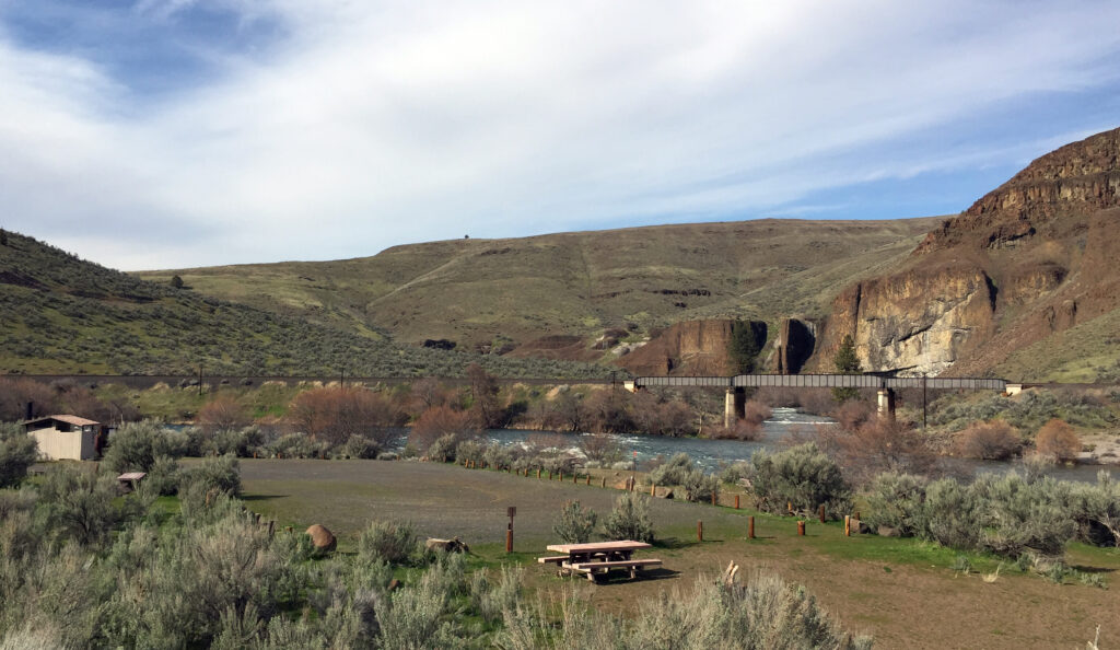 A serene landscape with a wooden picnic table in the foreground, lush green shrubs, a flowing river, and a rustic bridge against rolling hills.