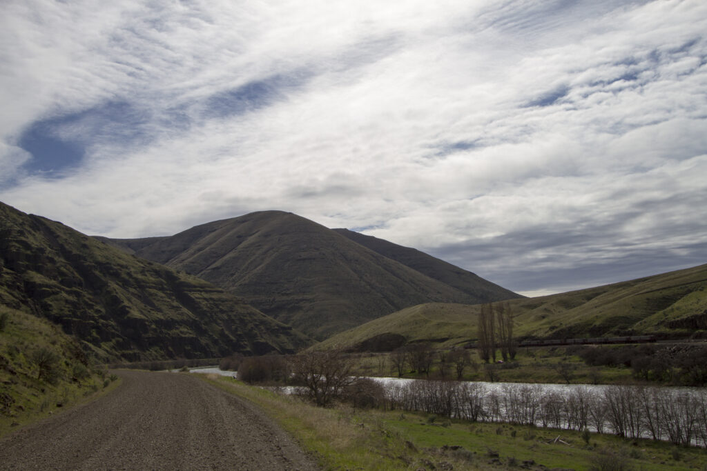 Gravel road alongside a river with gentle hills under cloudy skies. The scene has a serene, overcast ambiance with scattered bare trees.