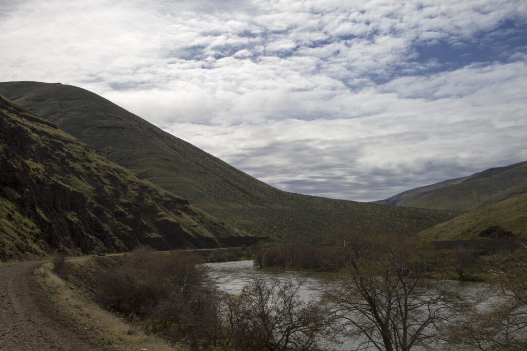 A winding river flows through a lush valley between two grassy hills under a partly cloudy sky. Sparse trees and a dirt path line the riverbank.
