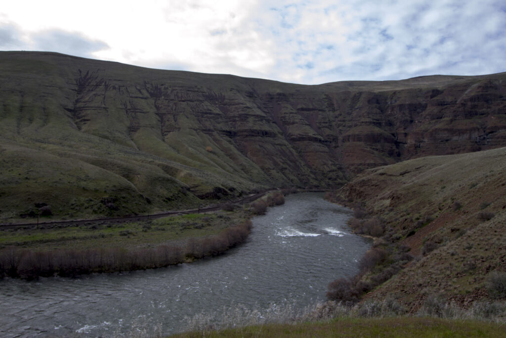 A winding river flows through a canyon with steep, grassy hills under a cloudy sky. The scene feels serene and remote, highlighting natural beauty.