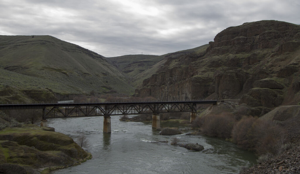 A steel railway bridge spans a calm river between rugged green hills and rocky cliffs under an overcast sky, creating a serene, expansive landscape.