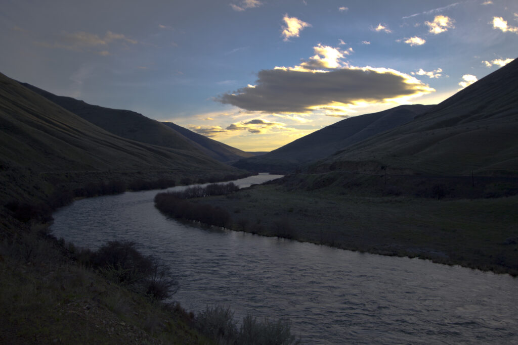 Sunset over a winding river between shadowy hills. The sky is vibrant with golden clouds, casting a serene and peaceful atmosphere.