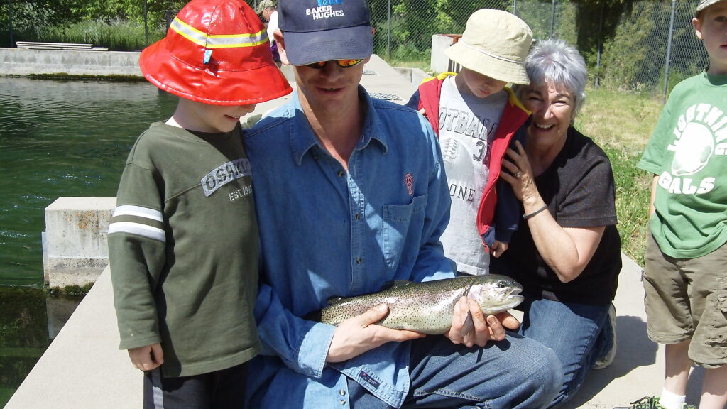 A person in a cap holds a large trout, surrounded by two children and an older woman smiling. The scene is outdoors by a pond, conveying joy.