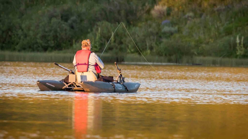 Man fishing in a small boat on a serene lake. He's wearing a red life vest, surrounded by lush greenery reflecting in golden water at sunset.