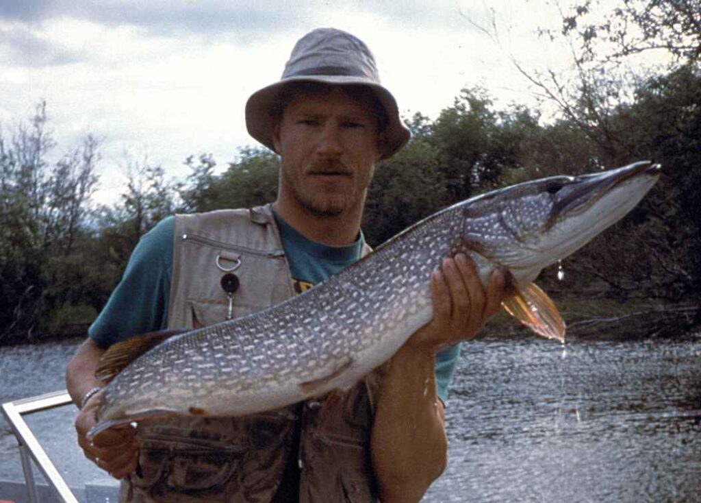A man in a fishing hat and vest proudly holds a large fish beside a river. The scene includes a boat with coolers and gear, set against a cloudy sky.