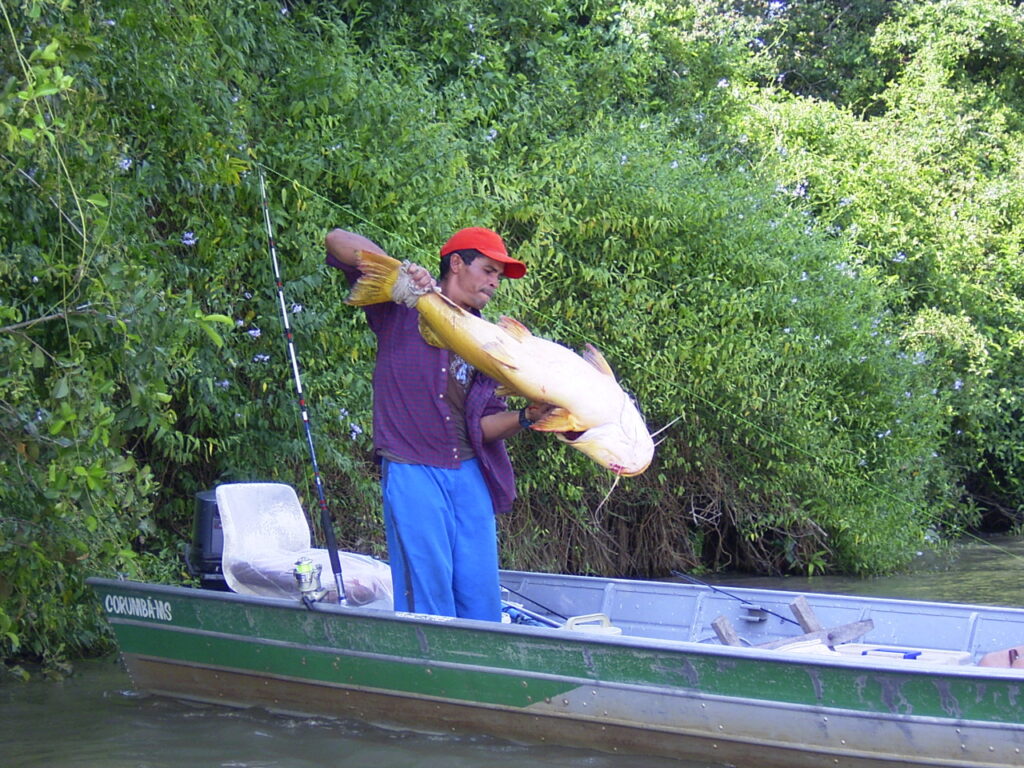 Man in a red cap and blue pants holding a large fish on a small green boat, surrounded by lush greenery, conveying a sense of excitement and achievement.