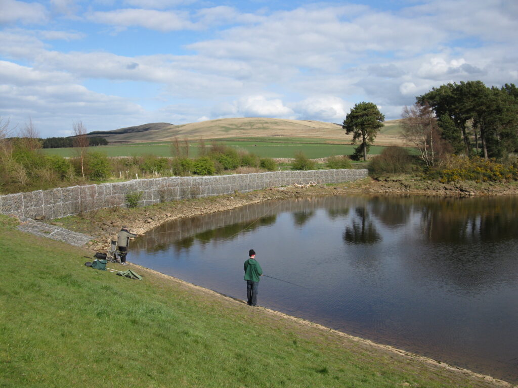 A serene pond with clear reflections, surrounded by lush green hills and sparse trees. Two people are fishing on the grassy bank, under a partly cloudy sky.