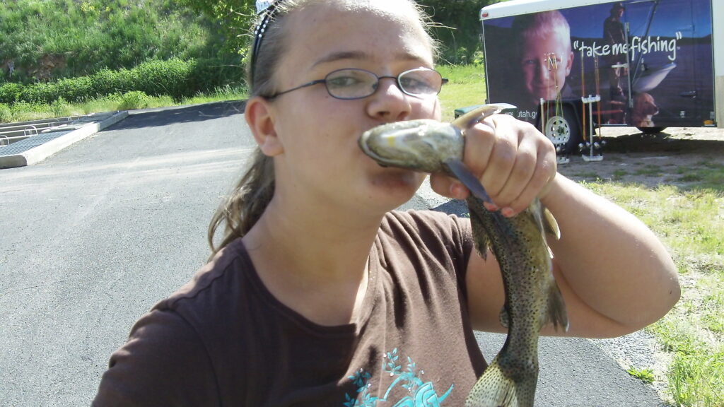 A person wearing glasses playfully kisses a fish they're holding outside. A trailer with "take me fishing" text is in the background. Bright, sunny day.