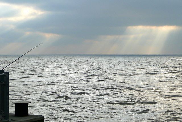 A fishing pole rests on a pier by a vast, rippling ocean under an overcast sky. Sunbeams break through clouds, casting a serene, contemplative mood.