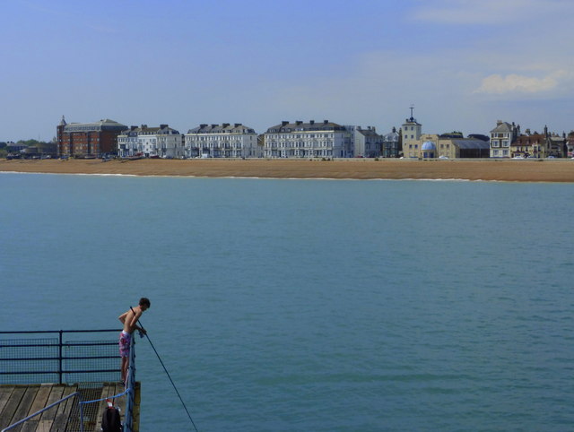 A person stands on a pier over calm waters, with a distant view of a coastal town and sandy beach under a clear blue sky. The scene is peaceful and serene.