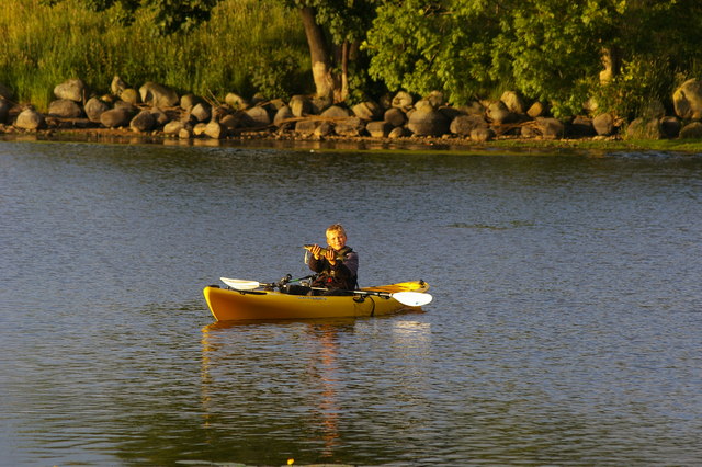 A person in a yellow kayak holds up a small fish, smiling on a calm lake with a rocky, tree-lined shore. The scene conveys a peaceful, joyful moment outdoors.