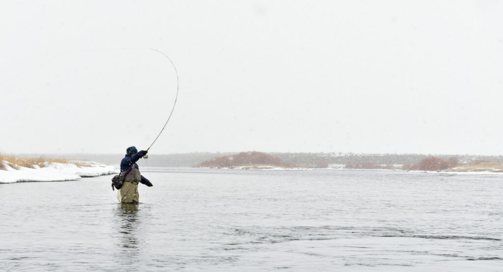 A person in waders fly-fishes in a snowy river under an overcast sky. Bare trees line the riverbank, creating a serene, wintry atmosphere.