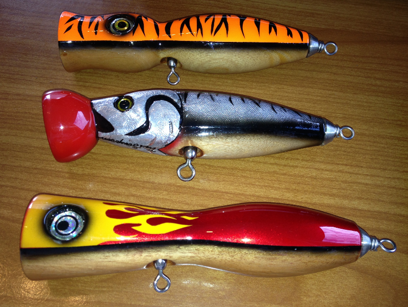 Three colorful fishing lures on a wooden surface. Top: orange with black stripes, middle: silver with red mouth, bottom: red with yellow flames.