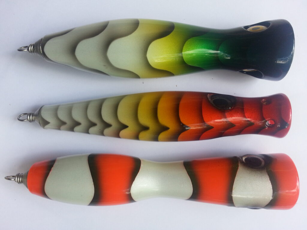 Three colorful fishing lures on a white background. Top is green-yellow, middle is orange-yellow, and bottom is red-orange. Each has a metal loop.