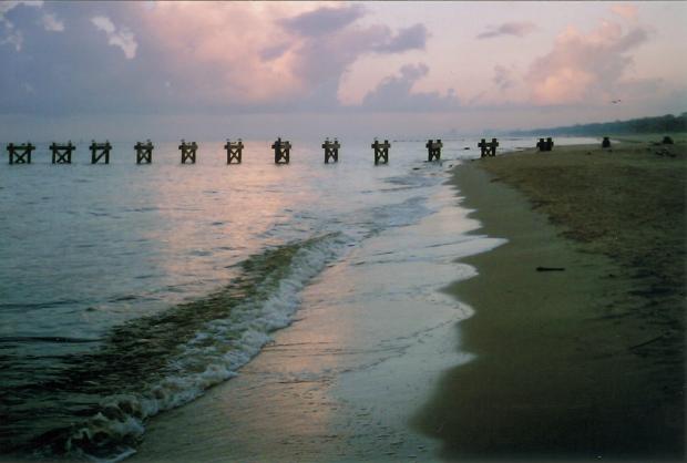 Serene beach scene at sunset with gentle waves, a sandy shoreline, and distant wooden pier remnants. Sky painted in soft pink and purple hues. Tranquil mood.