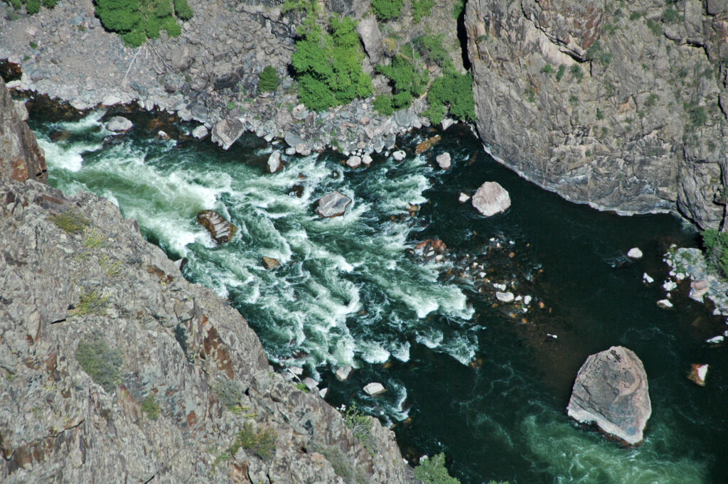 Aerial view of a rocky canyon with a turbulent river flowing through it. White water rapids contrast with dark green water, surrounded by rugged cliffs.