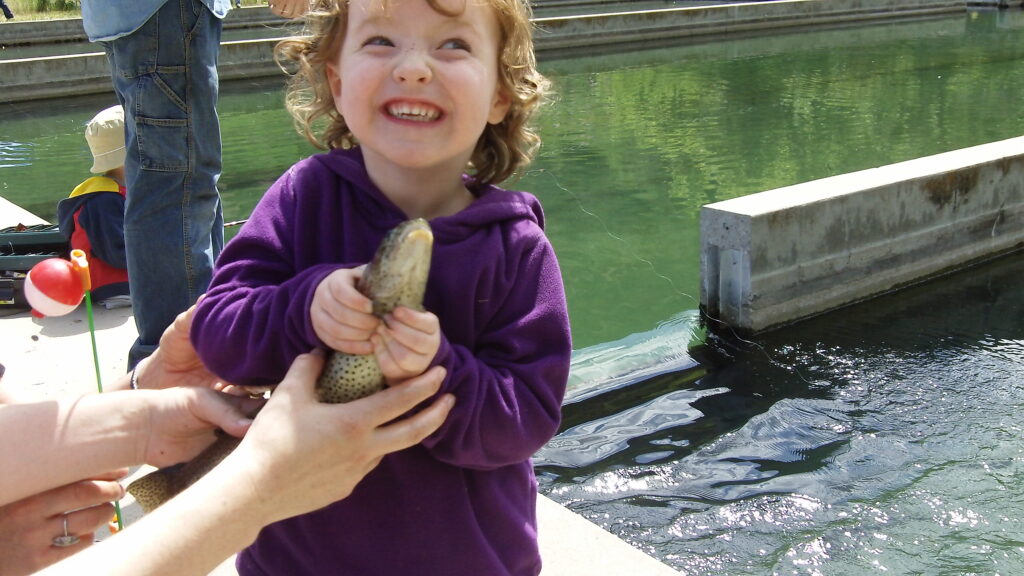A joyful child in a purple hoodie holds a fish with excitement near a pond, assisted by an adult's hands. The background is a calm, sunlit water scene.