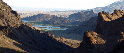 Rugged desert mountains frame a distant river winding through a sunlit valley, creating a dramatic, serene landscape with a sense of vastness.