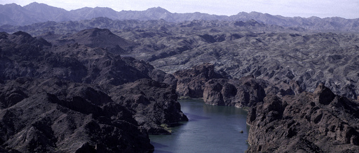 Aerial view of a serene river winding through rugged, rocky mountains. The landscape is vast and expansive, under a clear, pale sky, evoking tranquility.