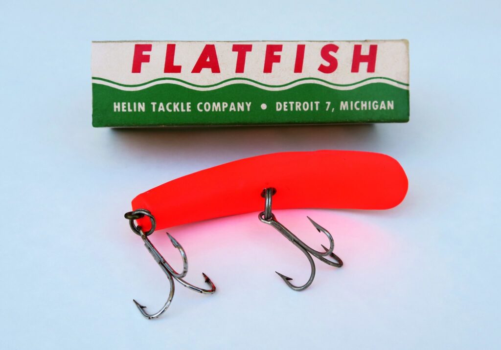 Bright orange fishing lure with two treble hooks beside a vintage "FLATFISH" box from Helin Tackle Company, Detroit, Michigan. Retro and vibrant.