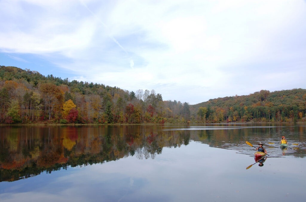 Two kayakers paddle on a calm lake reflecting autumn trees and a clear sky. The scene is serene, with vibrant fall colors and gentle ripples.
