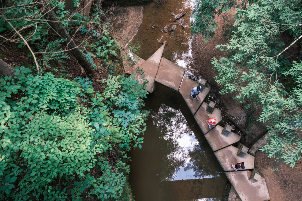 Aerial view of three people walking on a zigzag concrete path over a forest creek, surrounded by lush green trees and foliage, conveying a tranquil nature scene.