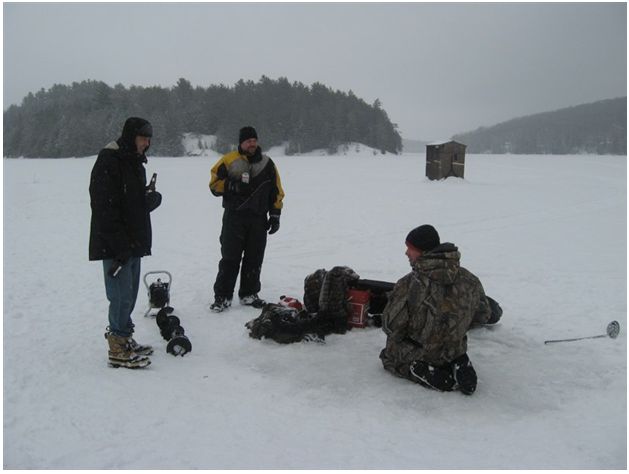 Three people on a snowy frozen lake appear to be preparing for ice fishing. One is seated, surrounded by gear. A small shack and forest are visible in the background, under an overcast sky, suggesting a cold, wintry atmosphere.