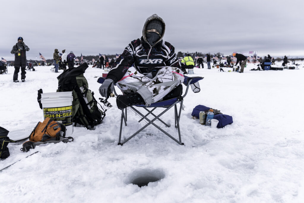 A person in winter gear ice fishing on a frozen lake, sitting on a chair over a fishing hole, surrounded by gear and a crowd in the background.