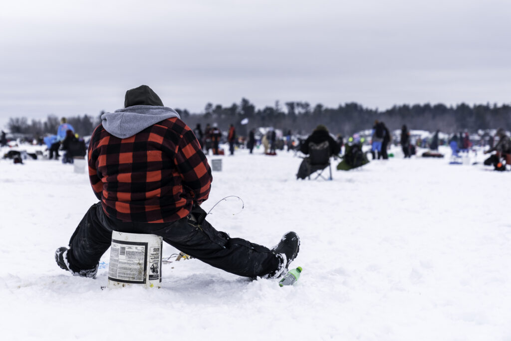 Person in a red plaid jacket sits on a bucket, ice fishing on a snow-covered lake. Others are scattered in the background, creating a communal, serene atmosphere.