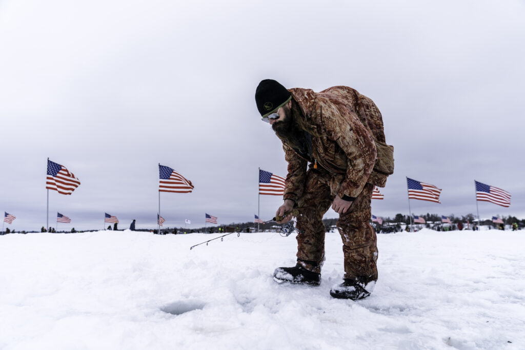 A person in winter gear ice fishes on a snowy field, bending over a hole. American flags wave in the background under a cloudy sky.