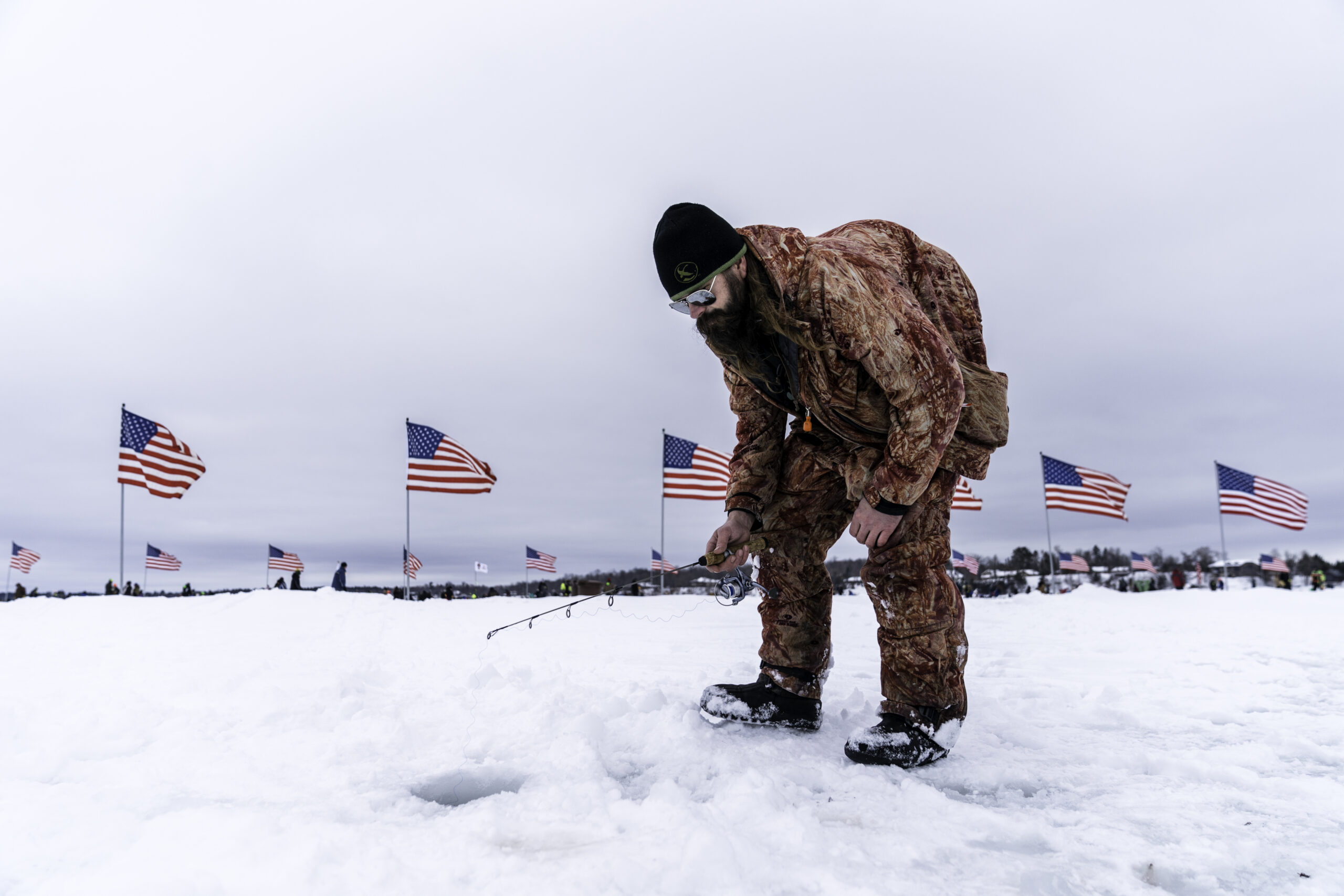 A person in winter gear ice fishes on a snowy field, bending over a hole. American flags wave in the background under a cloudy sky.