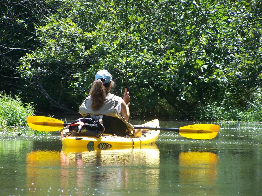 A person kayaking on a calm river, surrounded by lush green foliage. They wear a life vest and cap, holding a fishing rod, conveying a peaceful, adventurous vibe.