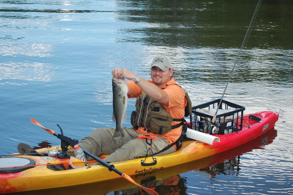 Man in an orange shirt and hat smiles while holding a large fish on a kayak. The calm water reflects the blue sky, creating a serene setting.