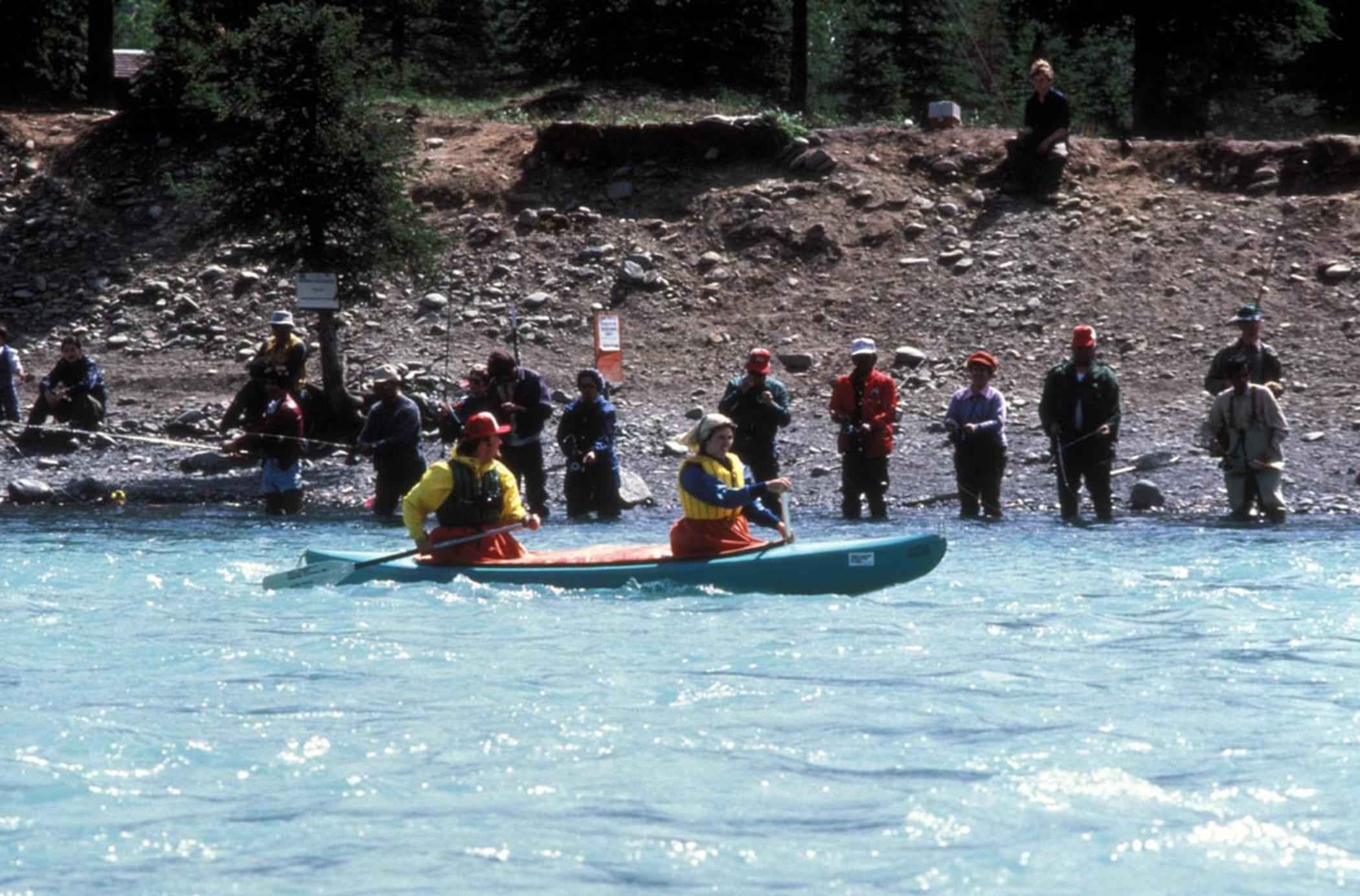 Two kayakers in colorful gear paddle through a turquoise river, watched by a group of people on the rocky shoreline, conveying a lively outdoor scene.