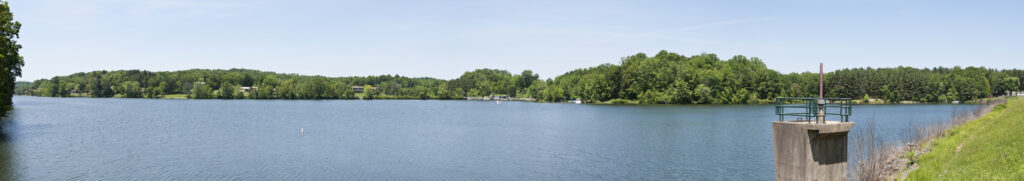 Panoramic view of a tranquil lake surrounded by lush green trees under a clear blue sky. A concrete platform extends into the water, adding a peaceful touch.