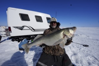 Man in winter gear proudly holds a large fish on a snowy landscape near an ice fishing trailer. Clear blue sky enhances the cold, victorious atmosphere.