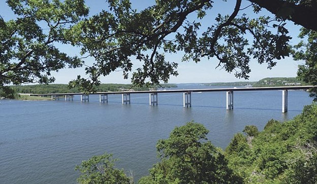 A long bridge stretches over a wide, calm lake, framed by lush green trees under a clear blue sky. The scene feels peaceful and expansive.
