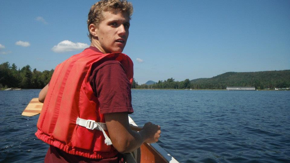 Young man in a red life vest paddling on a serene lake, surrounded by lush green hills under a clear blue sky. The scene feels peaceful and adventurous.