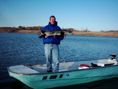 Man in blue jacket on a green boat proudly holds a large fish over calm waters, with a clear sky and distant autumn trees in the background.