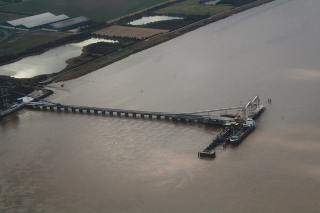 Aerial view of a large brown river with a long pier and a docked cargo ship. Surrounding industrial landscape with fields and warehouses visible.