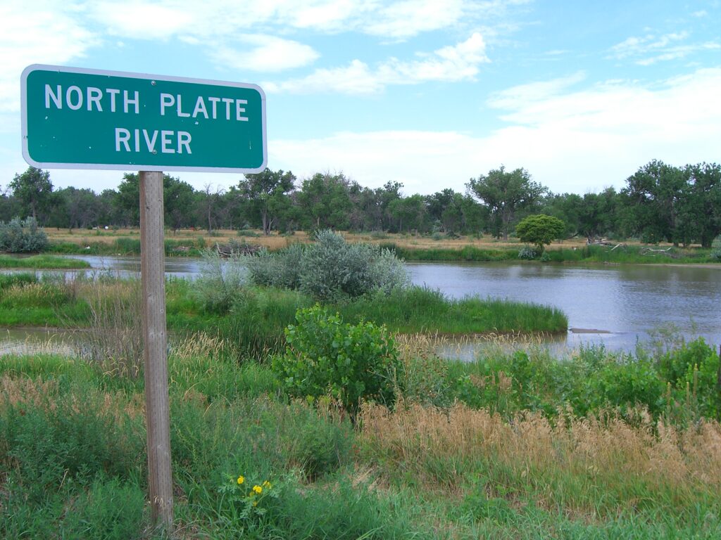 A green sign marked "North Platte River" stands beside a calm river, surrounded by lush greenery under a partly cloudy sky, conveying tranquility.